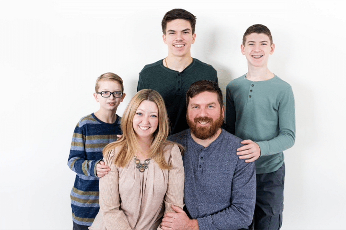 A family of five poses against a white background. A woman and a bearded man sit in front, smiling, while three boys stand behind them in casual long-sleeve shirts—ready for their next milestone or maybe searching for top hardwood floor refinishing nearby.