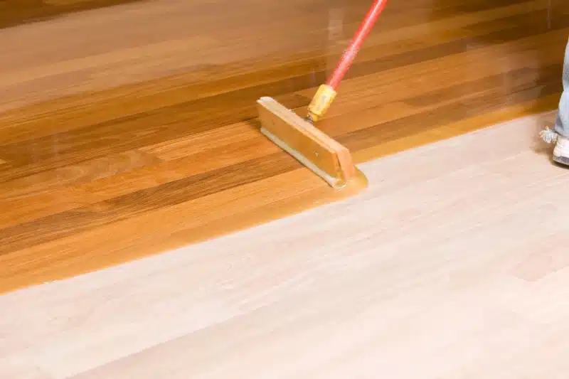 A person uses a mop to apply a clear finish to a wooden floor, showing the dramatic contrast achieved by professional hardwood floor refinishing, with one section glossy while the unfinished area remains lighter.