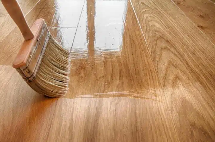 A close-up of a brush applying clear varnish to a wooden floor, highlighting the glossy, wet finish achieved through hardwood flooring and refinishing. A window reflection shimmers on the freshly polished surface.