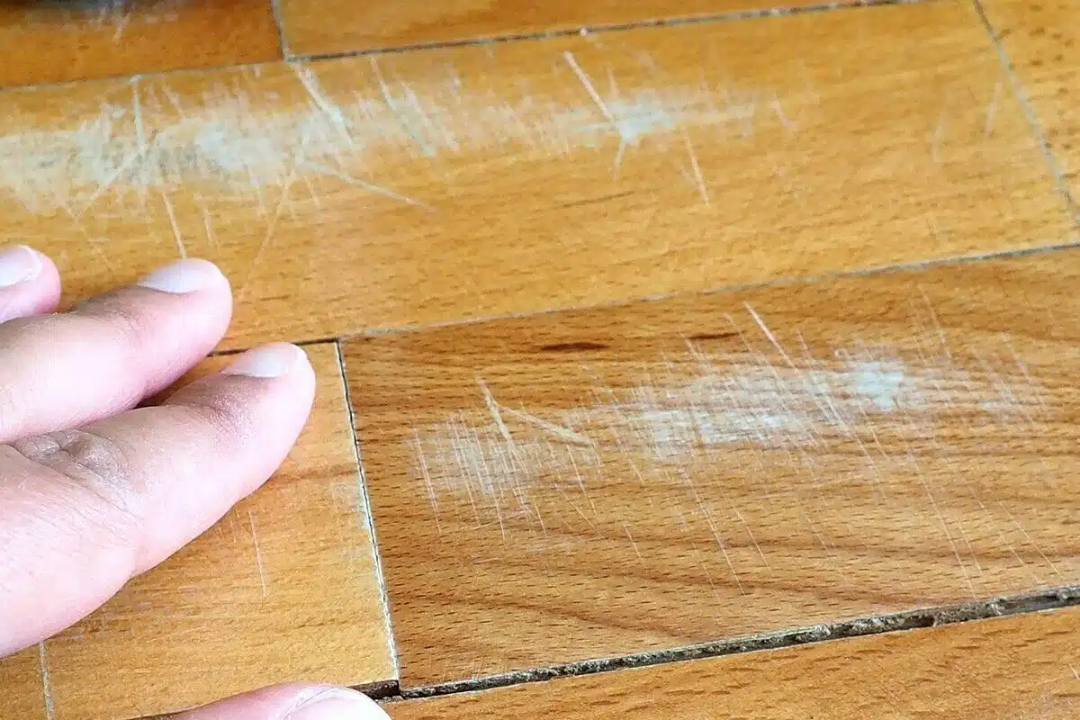 A hand touches a wooden floor with visible white scratches and scuff marks—perfect candidates for refinishing of hardwood floors to restore their original beauty.