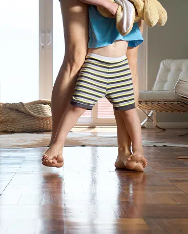 A small child in striped shorts stands on the feet of an adult, hugging their legs in a sunlit room with beautiful hardwood floors, showcasing the results of professional hardwood floor refinishing services—a basket rests in the background.