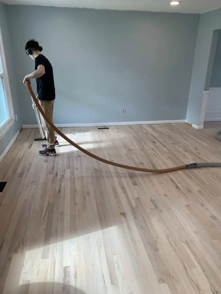a person wearing headphones vacuums dust from newly sanded hardwood floors in an empty room with light blue walls and a large window.