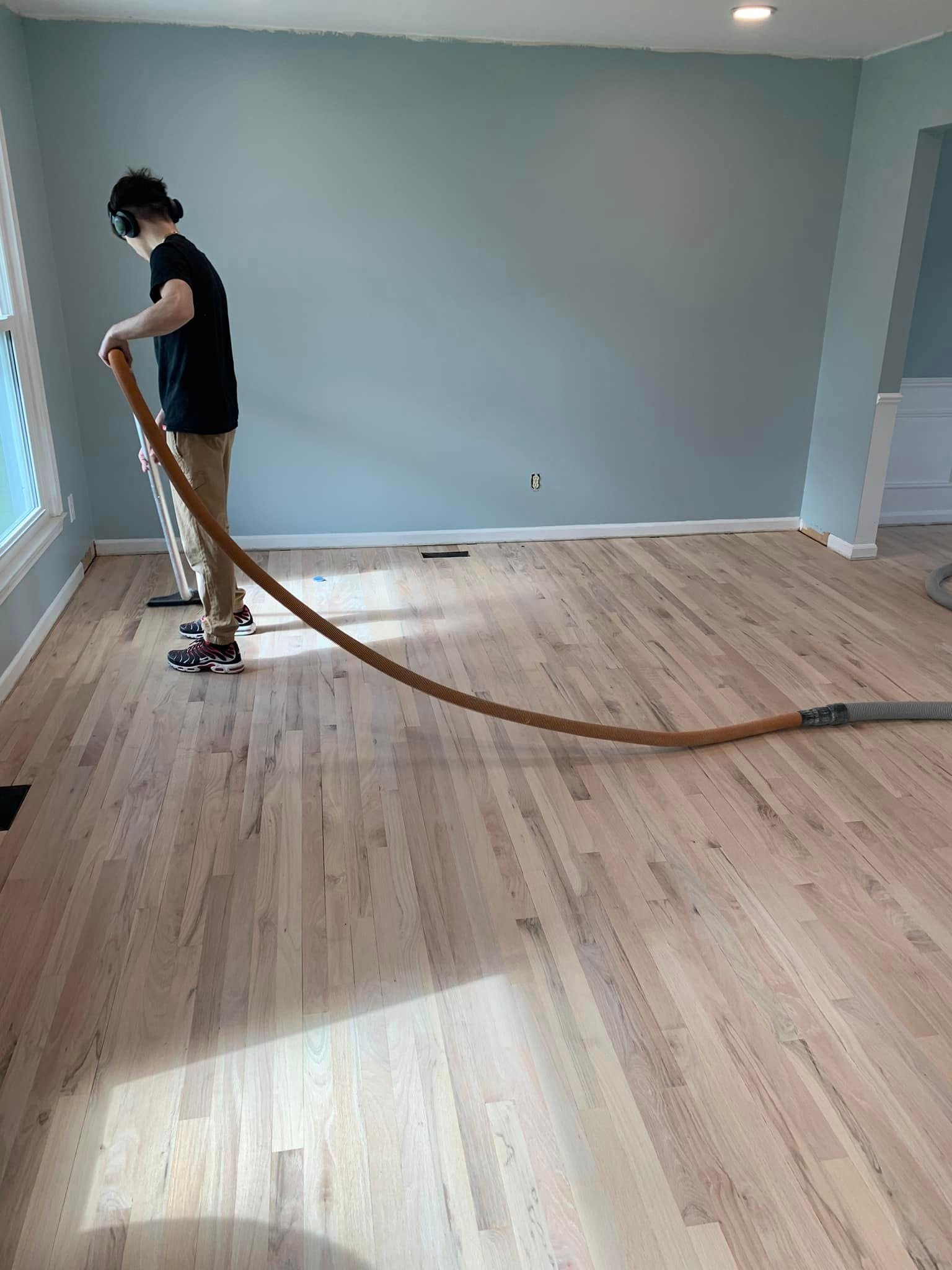 a person wearing headphones vacuums dust from newly sanded hardwood floors in an empty room with light blue walls and a large window.