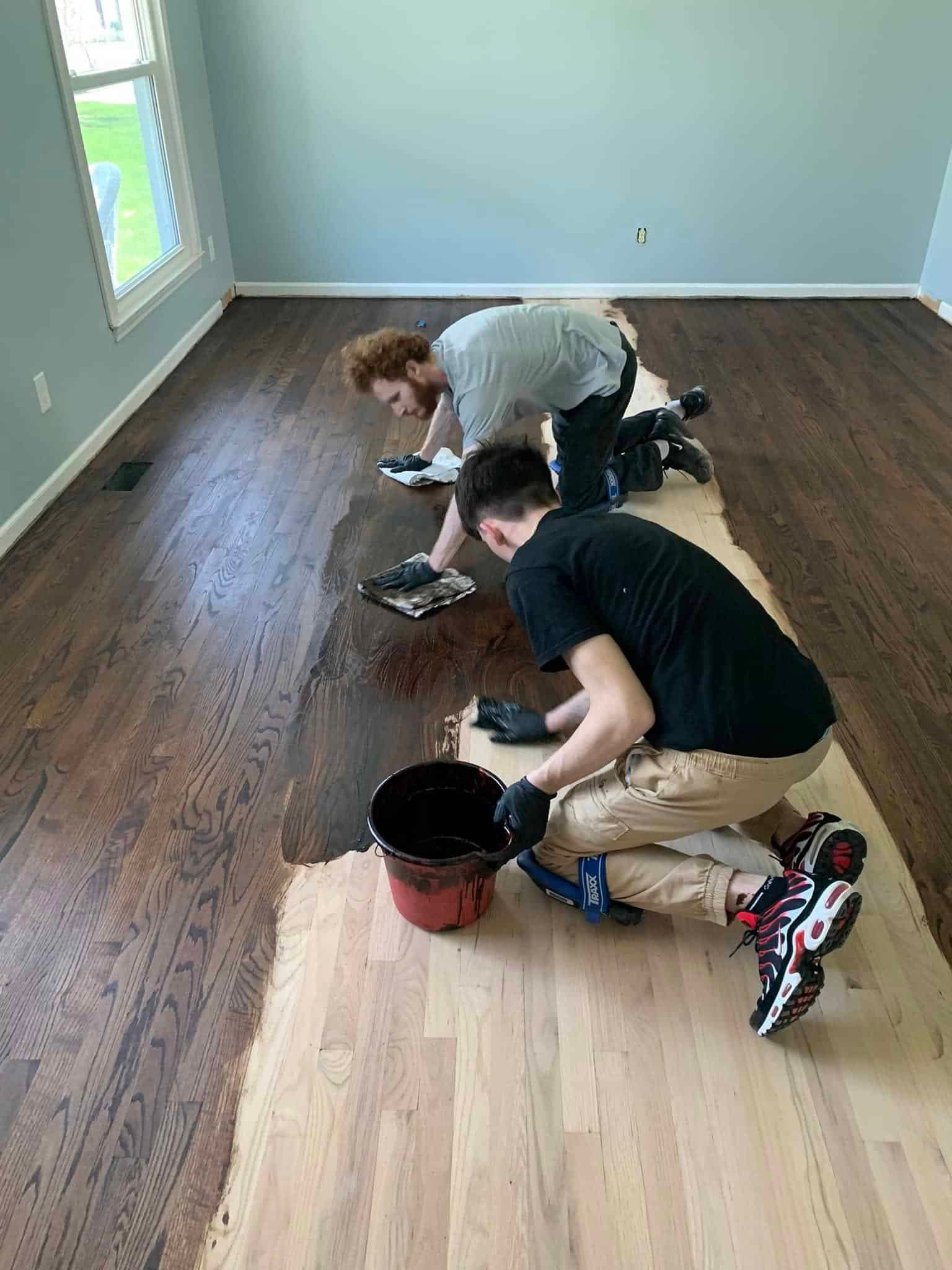 two people kneel and apply dark wood stain to a hardwood floor in a room with light blue walls, using rags and wearing gloves. a bucket of stain sits nearby, and much of the floor is still unfinished.