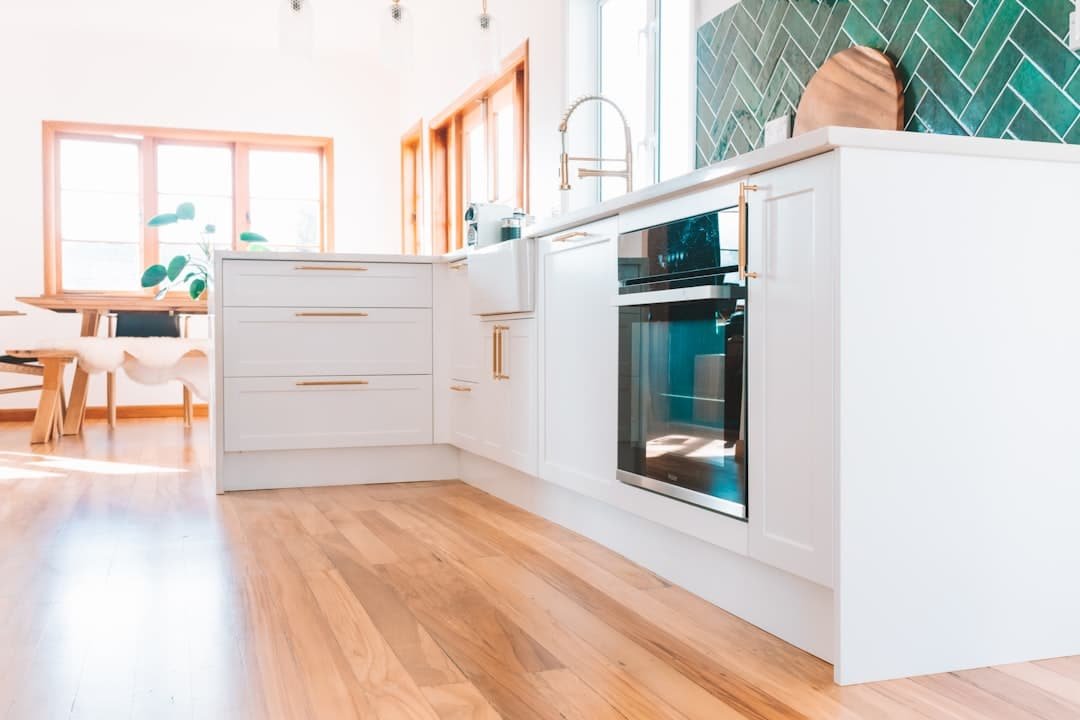 bright modern kitchen with white cabinets, gold handles, a built in oven, farmhouse sink, green tile backsplash, wooden floors, and large sunlit windows. dining area with a wooden table is visible in the background.